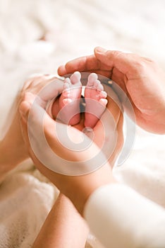 Mom`s and father hands are holding little cute legs of a newborn baby at home on a white bed.