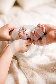 Mom`s and father hands are holding little cute legs of a newborn baby at home on a white bed.