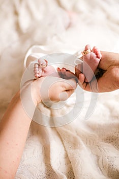 Mom`s and father hands are holding little cute legs of a newborn baby at home on a white bed.