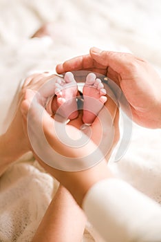 Mom`s and father hands are holding little cute legs of a newborn baby at home on a white bed.