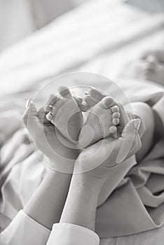 Mom`s hands are holding little cute legs of a newborn baby at home on a white bed