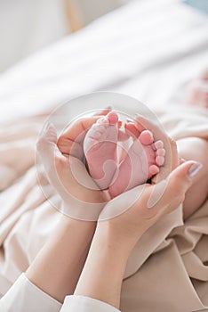 Mom`s hands are holding little cute legs of a newborn baby at home on a white bed