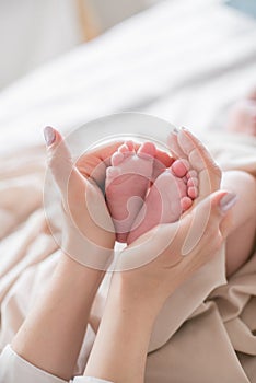 Mom`s hands are holding little cute legs of a newborn baby at home on a white bed