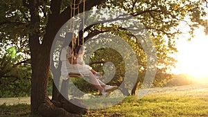 Mom shakes her daughter on swing under a tree in sun. mother and baby ride on a rope swing on an oak branch in forest