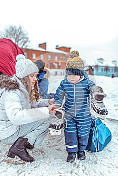 Mom shoes a child on skates to her son, a little boy of 3-5 years old, stands in a city park on a snow-covered skating