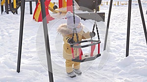 Mom shows curious newborn daughter a swing on a winter playground. The toddler is holding onto a baby swing and smiling.