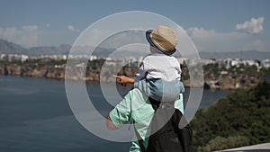 Mom shows toddler sitting on shoulders views of sea and town