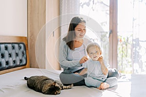 Mom sits with a little girl on the bed and combs her next to a lying tabby cat