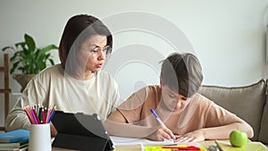 Mom, son doing homework and sitting at table with tablet in home room spbd.