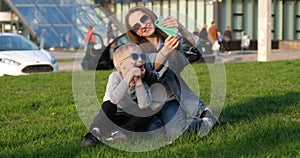 Mom and son laugh and take a selfie on a smartphone sitting on the lawn in the city. Mom and child have fun outdoors