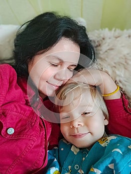 Mom and son lie on the bed, hugging each other and kissing. Mother with her son are having fun lying on top of each