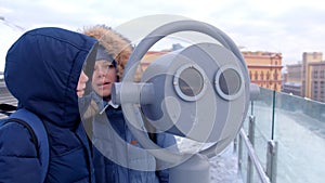 Mom and son looking through binoculars on roof of building in city in winter.