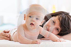 Mom and son lying down on bed in nursery room. Mother embracing infant baby. Kid looking at camera.