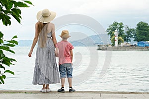 Mom and son standing on the pier on the sea background, lighthouse and mountains in the distance. Back view