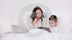 mom and son under blanket are sitting on soft bed in the hands of boy tablet playing online freelancer on laptop spend