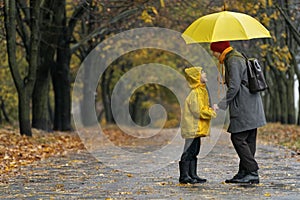 Mom and son are walking in the autumn park in rain with large yellow umbrella. Rainy day, back view