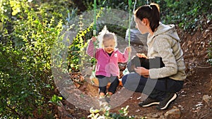 Mom squats and pets a dog on her lap next to a little girl on a swing