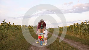 Mom teaches daughter to ride a bike on a country road in a field of sunflowers. a small child learns to ride a bike