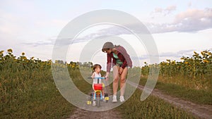 Mom teaches daughter to ride a bike on a country road in a field of sunflowers. a small child learns to ride a bike