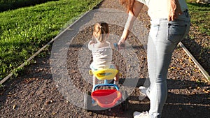 Mom teaches daughter to ride a bike in the park. A small child learns to ride a bicycle. Mother plays with her little