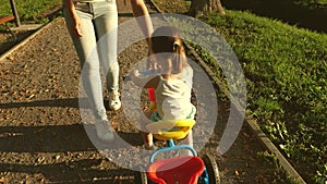 Mom teaches daughter to ride a bike in the park. A small child learns to ride a bicycle. Mother plays with her little