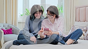 Mom and teenage daughter sitting on bed looking at smartphone, having fun laughing