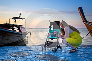 Mom with a toddler who is sitting in a stroller are standing on a pontoon pier waiting for a boat. Early morning, rising sky