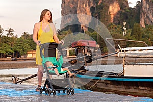 Mom with a toddler who is sitting in a stroller walks on a pontoon bridge against the background of boats. Sunrise light