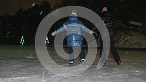 Mom together daughter skating on skates on night ice rink in winter park while snowfall. Mother and girl teenager