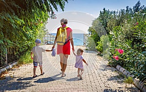 Mom and two children hold hands and walk along the path to the sea on clear, sunny summer day on vacation