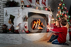 Mom and two little sons sit and relax under a Christmas tree by the fireplace,warm their hands by the fire.