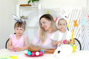 mom with two young daughters sits at a white table with a cake and a basket of Easter eggs and a white rabbit