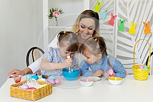 mom with two young daughters sits at a white table, painting Easter eggs.