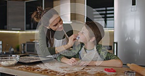 Mom wipes flour from her son's face in the kitchen. Happy mother and son cooking cookies together in the kitchen