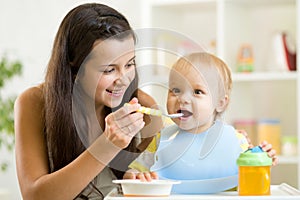 Mommy giving healthy food to baby son on high chair in kitchen.