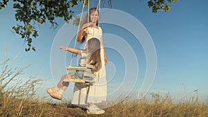 Mommy shakes her healthy little daughter on a swing under a tree in sun. free family is having fun in park.mother plays