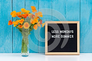 Less mondays more summer. Motivational quote on letter board and bouquet orange flowers on white table against blue wooden wall.