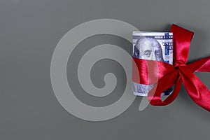 Money as gift concept, win or bonus, hundred dollar bills on grey background. Rolled 100 dollar bills is tied with red ribbon with