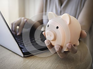 Money box on hand. Hand holding the pink piggy bank while using laptop computer on desk.