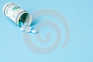 Money dollar rolled up with pills flowing out  on blue background, high costs of expensive medication concept. Copy space