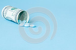 Money dollar rolled up with pills flowing out isolated on blue background, high costs of expensive medication concept. Copy space