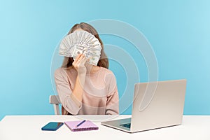 Money making advertisement. Woman employee sitting at workplace and covering her face with dollar banknotes