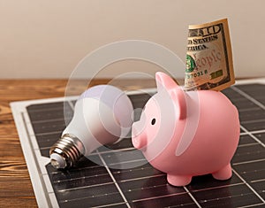 Money, a piggy bank, a LED lamp and a house model on the background of solar panels, close-up.