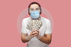 Money. Portrait of suprised young hipster man with surgical medical mask in white shirt and casual hat, stand, holding fan of