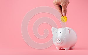 Money saving and investment concept. Hand putting Bitcoin coin to piggy bank on pink background with copy space