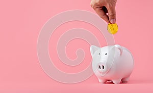 Money saving and investment concept. Hand putting Bitcoin coin to piggy bank on pink background with copy space