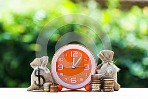 Money bags and orange clock on wooden table with nature backgro