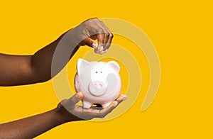 Money Savings. Unrecognizable black woman putting coin into piggybank over yellow background