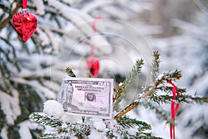 Money in us dollars on a Christmas tree with red heart decorations, copy space. New Year in a winter park with snowy trees