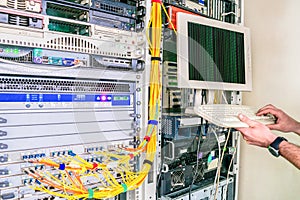 The monitor and keyboard are in the server room. Male hands on the keyboard. The programmer stands next to the powerful servers